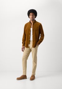 Bruine corduroy knoopshirt over een witte tanktop, gecombineerd met beige broek en bruine suède instappers, staand voor een effen achtergrond.