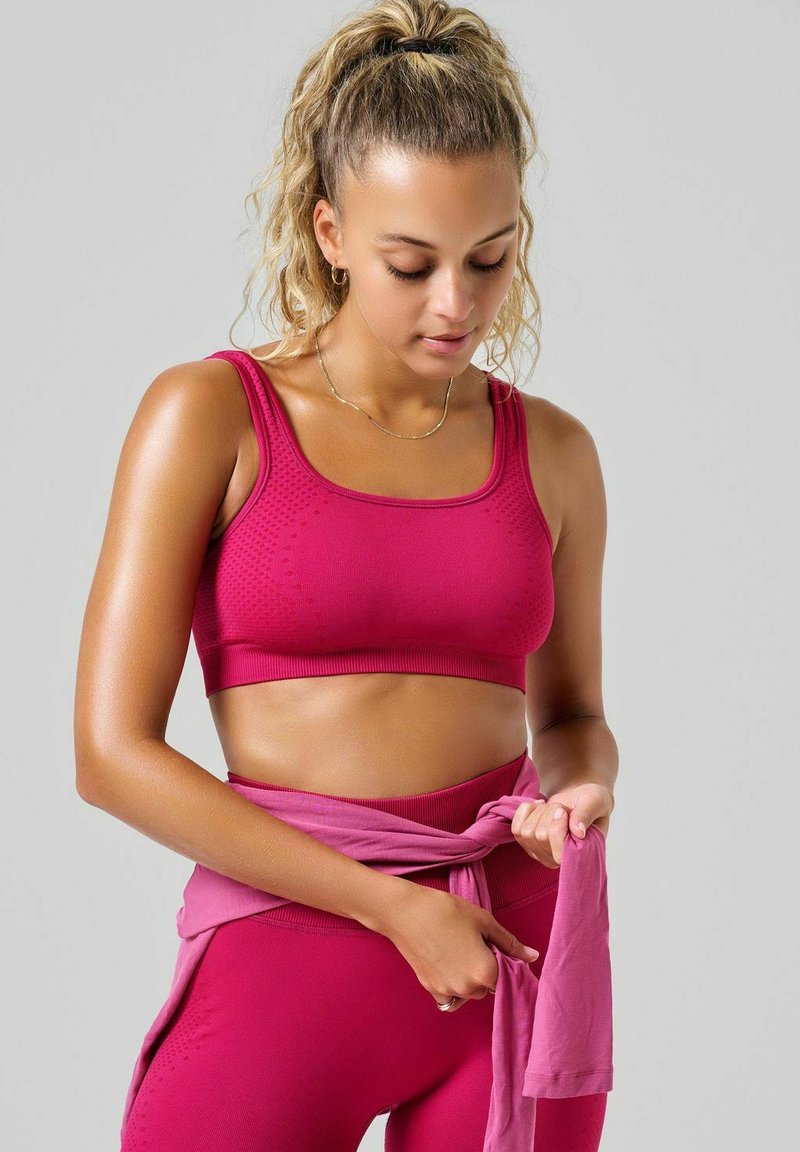 Dopasowany sportowy crop top i legginsy wysoki stan w jednolitym ciemnoróżowym kolorze. Top ma szerokie ramiączka i teksturalne detale, a materiał wydaje się elastyczny.
