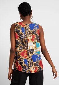 Ärmlös topp med ett färgglatt, mönstrat tyg som har röda, blå och svarta sektioner, guldaccenter och prickar. Slät textur.