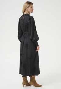 Musta pitkähihainen mekko, jossa on tekstiilinen kukkakuosi, joustava vyötärö ja avainaukko selkädetaili, yhdistettynä beigeihin nilkkureihin.