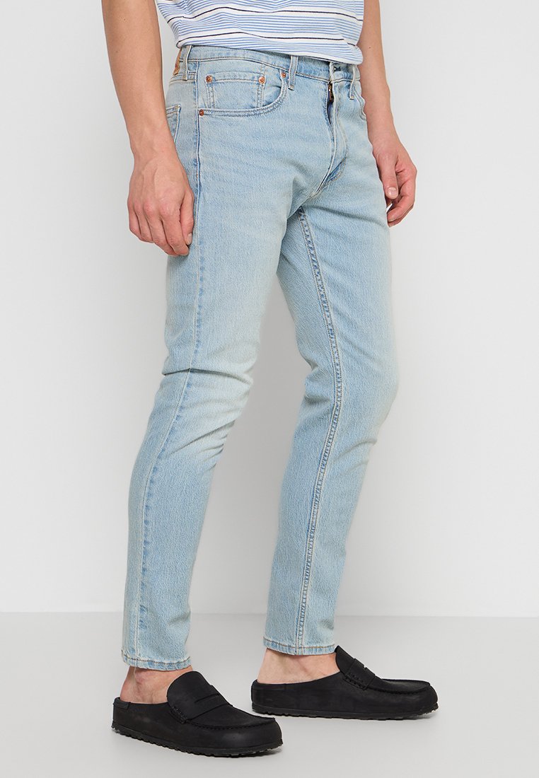Jeans en denim bleu clair avec une coupe slim, présentant un design classique à cinq poches et une légère décoloration, associés à des chaussures noires à enfiler.