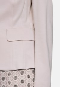 Helles rosa Blazer mit glatter Textur, ausgestattet mit einer Klappentasche und einer strukturierten Passform. Kombiniert mit einem gemusterten Rock mit kreisförmigen Motiven in Grau.