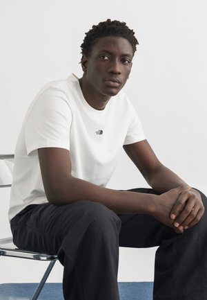 Tânăr așezat pe un scaun metalic, purtând un tricou alb The North Face și pantaloni negri, cu mâinile împreunate, privirea ușor spre o parte.