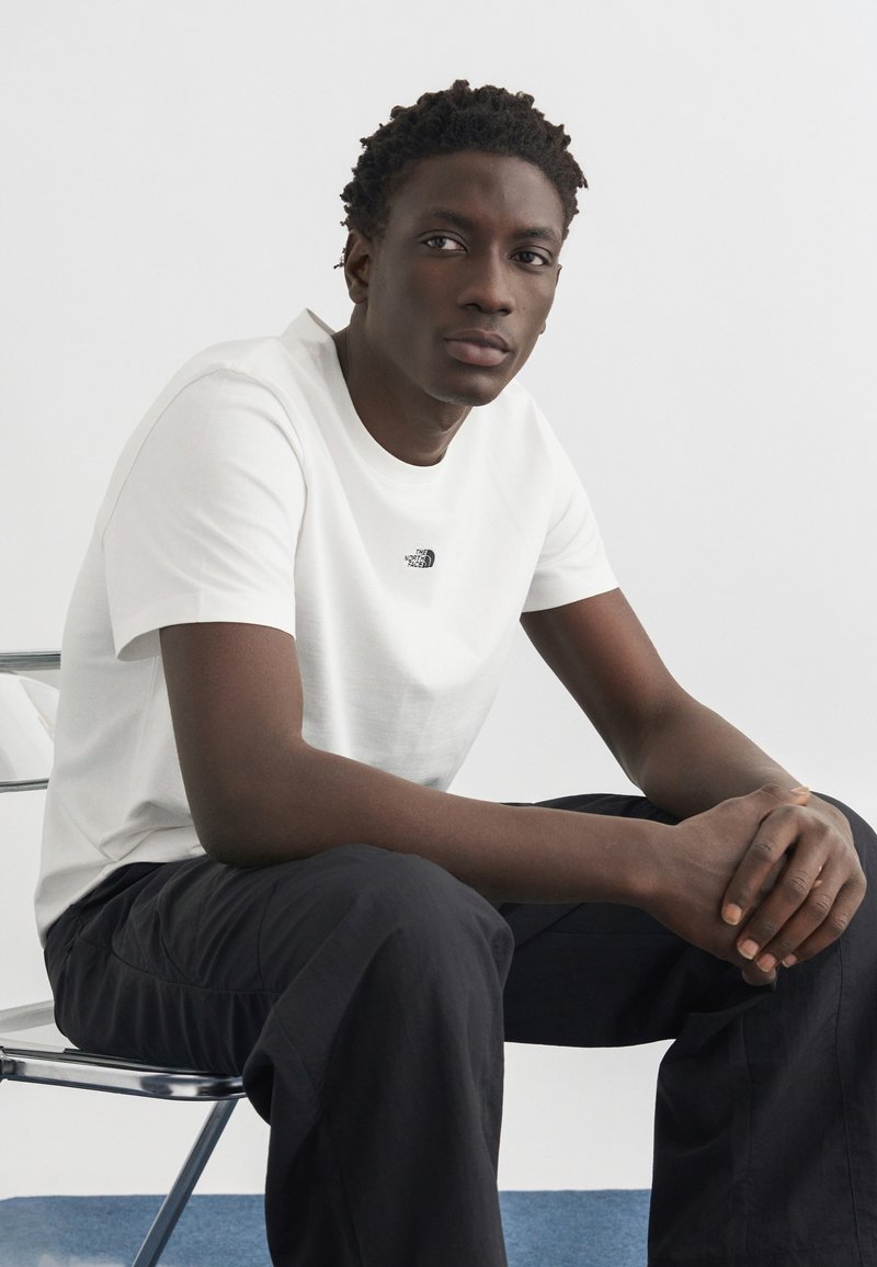 Νεαρός άνδρας καθισμένος σε μεταλλική καρέκλα, με λευκή μπλούζα The North Face και μαύρο παντελόνι, χέρια σταυρωμένα, κοιτάζοντας λίγο προς τα πλάγια.