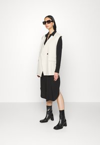 Colete sem mangas em marfim sobre um vestido preto com botões, combinado com botas de salto alto pretas até o joelho. O colete apresenta bolsos e um detalhe com botão.