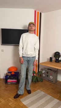 Gray sweatshirt, light blue jeans, dark shoes. Standing against a wall with colorful stripes, featuring a TV and modern decor.