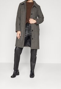 Donna che indossa un lungo cappotto grigio tartan, un maglione marrone, pantaloni in pelle nera e stivaletti neri su uno sfondo chiaro e semplice.