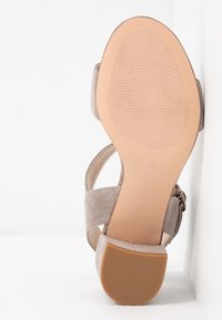Sandale en daim beige avec un talon bloc, une semelle texturée et une bride à la cheville, vue du dessous sur un fond blanc.