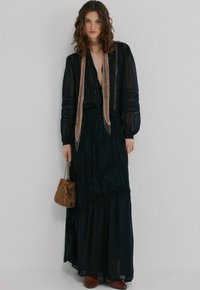 Robe longue noire en tissu transparent, avec des volants superposés, des détails brodés et un décolleté plongeant en V. Assortie d'un sac à main marron à motifs et d'une écharpe légère.