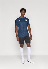 Maglia da calcio blu navy con righe textured, dettagli bianchi e blu, abbinata a pantaloni corti neri e calze bianche, con un logo sul petto.