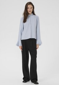 Žena s dlouhými hnědými vlasy, oblečená v lehké modré halence s vysokým výstřihem a rozšířenými rukávy, v volných černých kalhotách, stojící na čistém bílém pozadí.