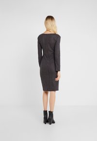 Donna con lunghi capelli biondi, che indossa un vestito aderente a maniche lunghe e lungo fino al ginocchio a quadri, e stivaletti neri con tacco, in piedi con la schiena rivolta verso di noi.