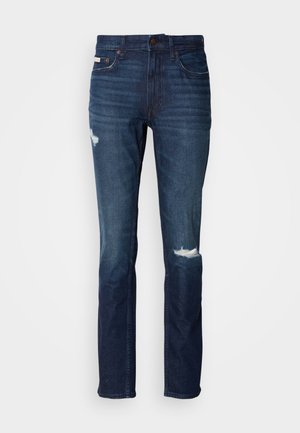 Jean en denim bleu foncé avec une coupe slim, présentant deux patchs usés à l'avant, des coutures robustes et un design classique à cinq poches.