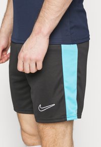 Mustat urheilushortsit, joissa on tekstuuriinen vaaleansininen sivunauha ja valkoinen Nike-logo alhaalla edessä.