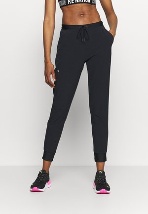 Žena nosící černé tepláky Under Armour, černou sportovní podprsenku s bílým logem a černé tenisky Puma s růžovými podrážkami stojící na bílém pozadí.
