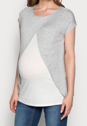 Gravid kvinde iført en tofarvet graviditetstop med et lys gråt overlay og en hvid underlayer, kombineret med blå jeans.