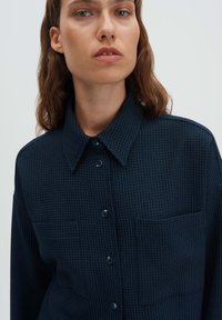 Navy blue textured shirt with a pointed collar, button front, and two chest pockets. Features a patterned fabric with a subtle grid design.