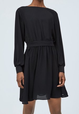 Žena stojící před jednobarevným světlým pozadím, oblečená v černých dlouhých rukávech s obtáhnutým pasem a mírně rozšířenou sukní.