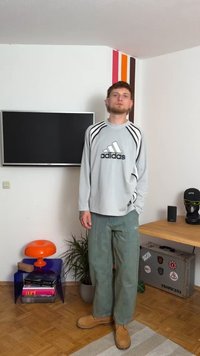 Camisa larga de manga gris de malla de Adidas con rayas negras, pantalones holgados verdes y botas de ante color beige. El fondo incluye un televisor y una decoración colorida.