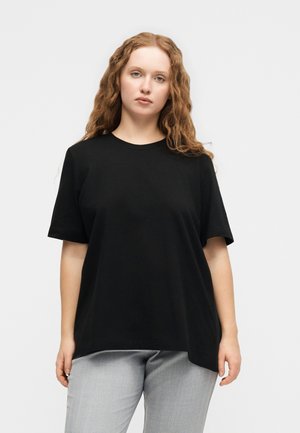 Jauna moteris su ilgais garbanotais plaukais, dėvinti laisvą juodą marškinėlį ir šviesiai pilkas kelnes, stovinti priešais paprastą šviesų foną.