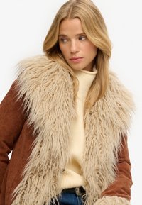 Brun suedepels med en fluffy, lys beige faux fur-krave. Det indre lag er en cremet strik. Texturn forskellene mellem materialerne.