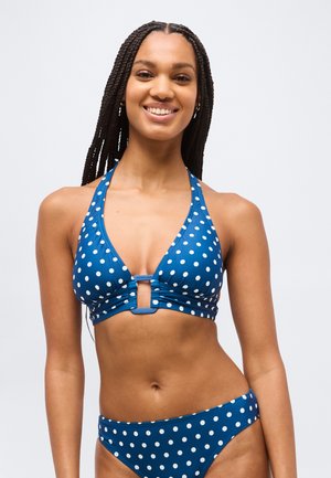 Femme souriante aux longues tresses portant un haut de bikini licou bleu et un bas assorti à pois blancs sur un fond uni.