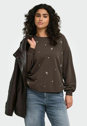 Kvinde med krøllet mørkt hår iført en brun sweatshirt med små hvide prikker og lyseblå jeans, der holder en mørk læderjakke over den ene skulder.