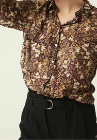 Blusa floreale marrone trasparente con fiori bianchi e viola, design con bottoni frontali e vestibilità comoda. Pantaloni neri a vita alta con cintura.