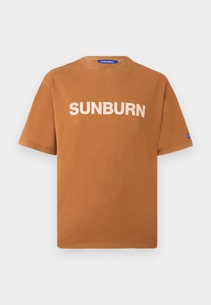 Brązowy T-shirt z krótkim rękawem z białym napisem "SUNBURN" z przodu i małą niebieską metką na lewym rękawie, wyświetlony na jednolitym tle.