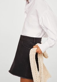 Falda de mezclilla negra en forma de A con dobladillo deshilachado, combinada con una blusa blanca de manga larga y sosteniendo un accesorio de punto de color claro.