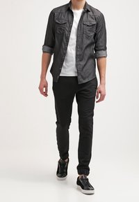 Ciemnoszara koszula z guzikami na białym T-shircie, w zestawieniu z czarnymi tapered spodniami i czarnymi trampkami, o gładkiej fakturze i podwiniętych rękawach.