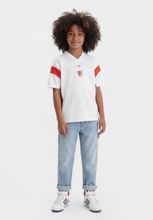 Gutt med krøllete hår som har på seg en hvit sportsskjorte med røde striper, lyseblå oppbrettede jeans og hvite joggesko med røde og blå detaljer.