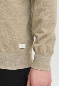 Beige gebreide trui met ribgebreide manchetten en zoom, voorzien van een stoffen label met tekst aan de onderlinkerkant. Zachte textuur en casual ontwerp.