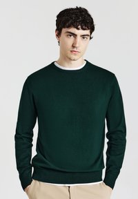 Dunkelgrüner Strickpullover mit Rundhalsausschnitt, gerippten Ärmelbündchen und Saum. Über einem weißen T-Shirt getragen, das eine glatte Textur und eine figurbetonte Passform aufweist.