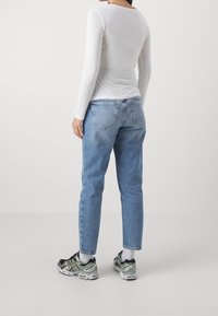 Vit långärmad topp, ljusblå raka jeans, svarta och grå sneakers med reflektiva detaljer, sedd bakifrån mot en neutral bakgrund.