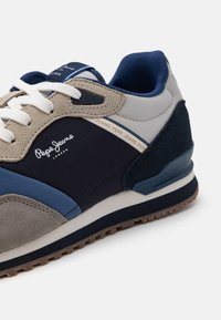 Navy and tan sneaker with suede and mesh materials. Features white laces, blue accents, adhesive sole, and brand logo on the side.