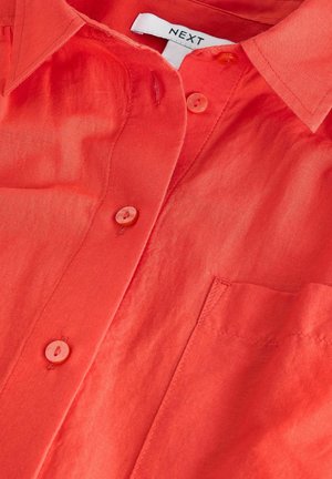 Camisa corta de manga corta, roja y con botones, hecha de una tela ligera. Presenta un bolsillo en el pecho y botones a juego. La etiqueta dice "NEXT."
