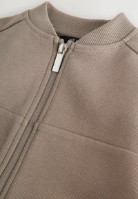 Veste zippée beige avec col côtelé et tissu texturé. Présente une fermeture éclair argentée et deux poches plaquées à l'avant. Design simple et détails de couture.
