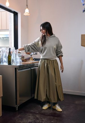 Kobieta w szarej bluzie z kapturem i oliwkowej spódnicy sięga po szklankę stojącą na blacie z butelkami i ciastem, znajdując się w nowoczesnej kuchni.
