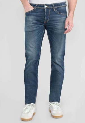 Jeans in denim blu slim-fit con leggero sbiadimento, design a cinque tasche e chiusura con bottone, abbinati a sneakers bianche con suola in gomma.