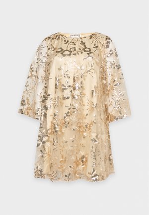Une femme porte une robe droite couleur crème, légèrement transparente, ornée de broderies florales dorées et de manches évasées, avec des sandales beiges à talons hauts, sur un fond uni.