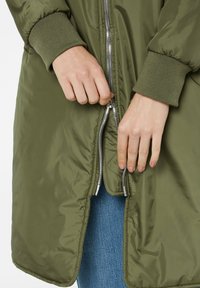 Olivgrüne Zip-Jacke mit glänzendem Reißverschluss. Verfügt über gerippte Bündchen und eine glatte, leicht reflektierende Textur. Die Hände sind zu sehen, wie sie den Reißverschluss anpassen.