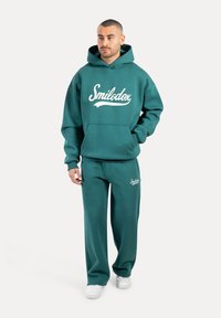 Sudadera de algodón color aguamarina con el logo blanco de "Smilodox", que cuenta con un bolsillo frontal y capucha con cordón, combinada con pantalones de chándal a juego en color aguamarina.