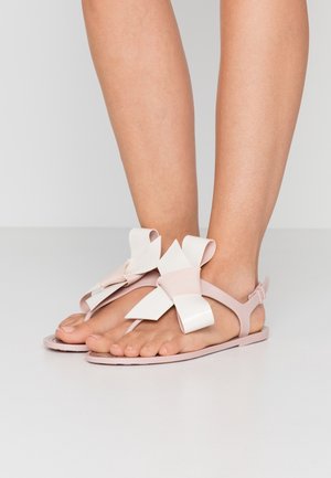 Des jambes portant des sandales plates rose pâle avec de grands nœuds décoratifs sur le dessus des pieds, se tenant sur une surface blanche lisse.