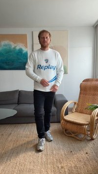 Sudadera blanca con logo azul de "Replay", jeans negros y zapatillas grises. Entorno interior con un sofá gris y una silla de mimbre. Luz natural.