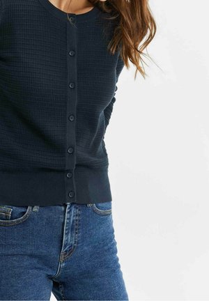 Femme portant un cardigan boutonné texturé bleu marine et un jean en denim bleu sur un fond clair uni.