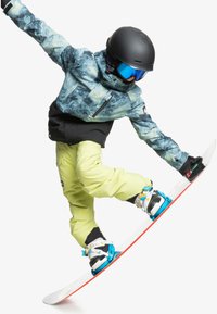 Snöbrädaåkare i en marmorblå och grön jacka, svarta byxor och ljusgula snöbyxor. Bär en svart hjälm och blå goggles, utför ett hopp.