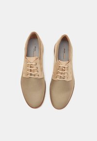 Scarpe beige con lacci, realizzate in mesh e suede, punta rotonda e suola marrone a contrasto. Presentano lacci piatti e un design minimalista.