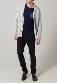 Světlý šedý cardigan s šálovým límcem a tmavými knoflíky, nošený přes námořnicky modrou košili s kapsou, doplněný černými džínami a tmavými botami.