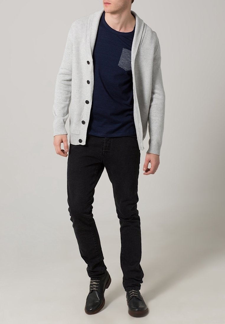 Světlý šedý cardigan s šálovým límcem a tmavými knoflíky, nošený přes námořnicky modrou košili s kapsou, doplněný černými džínami a tmavými botami.